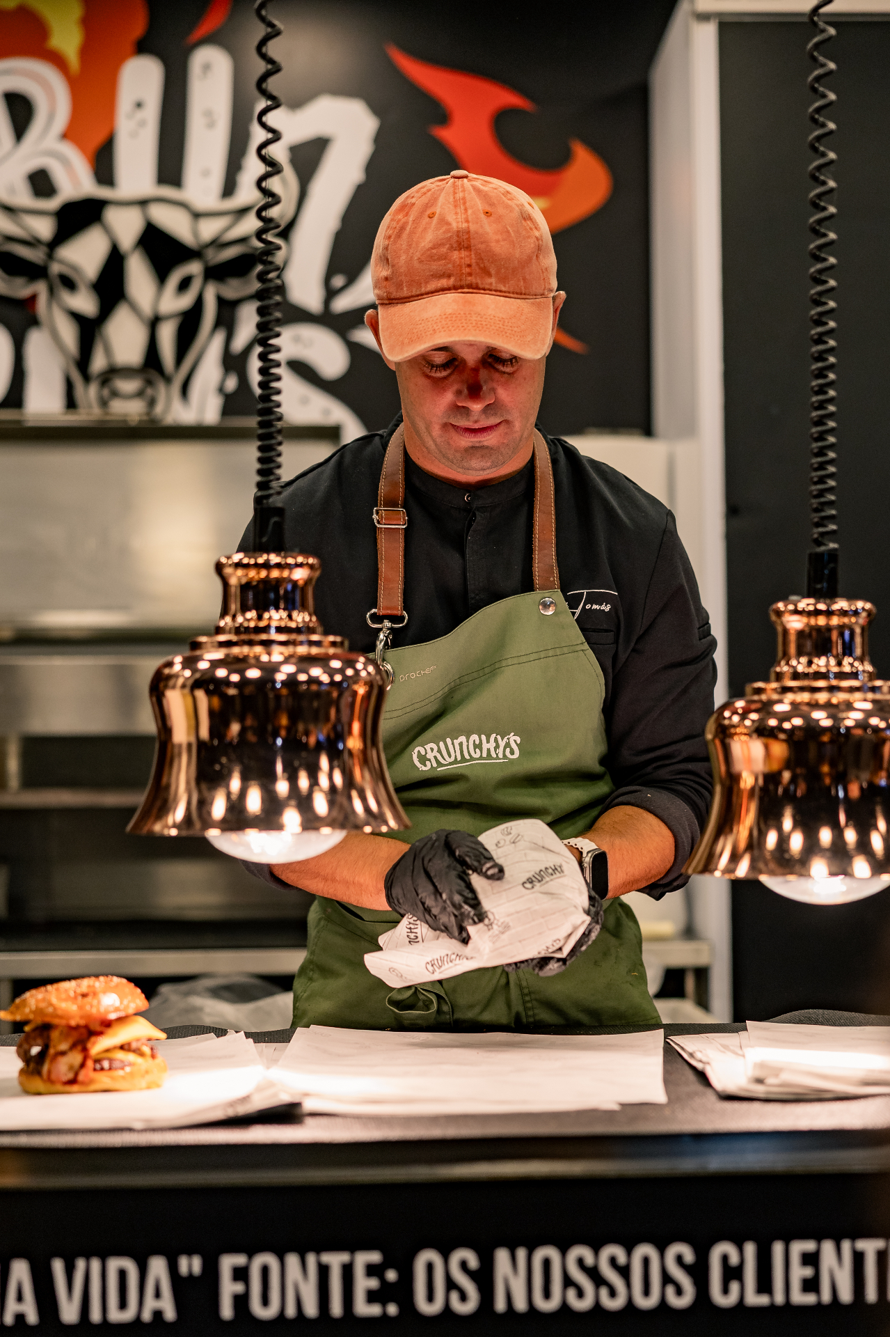 Membro da equipa Crunchy's, de avental verde com logótipo, a empacotar um hambúrguer sob lâmpadas de cobre